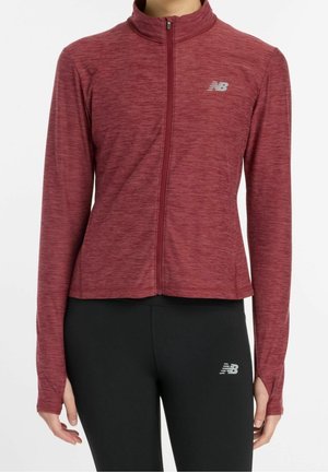 Woman wearing a maroon full-zip athletic jacket with thumb holes and black fitted leggings with logos on the chest and thigh.