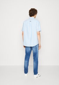 Camicia a maniche corte con bottoni di colore azzurro chiaro e righe verticali bianche; presenta un colletto a punta e un gancio posteriore per appenderla. Indossata con jeans blu.