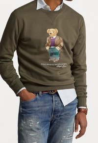 Sudadera de color verde oliva con una ilustración de un oso con blazer, vistiendo jeans azules y una camisa de rayas claras visible.
