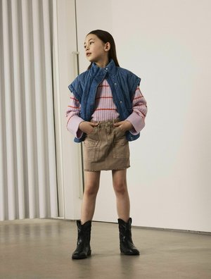 Gilet en denim matelassé, pull rayé rose et rouge, jupe en denim beige avec un ourlet effiloché, et bottes de cowboy noires. Look simple et superposé.