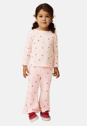 Ragazza giovane con capelli castani a code di cavallo che indossa un top e pantaloni coordinati rosa chiaro a coste con piccole stampe di fiori rossi e sneakers rosse.