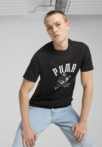 Juodas trumpas rankovėmis marškinėliai su baltu "PUMA" logotipo grafiniu printu, derinami su šviesiai mėlynomis džinsinėmis kelnėmis. Lygus paviršius ir laisvas siluetas.