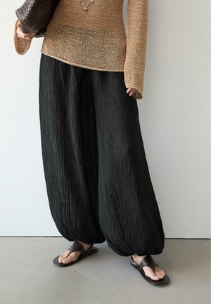 Osoba nosząca luźne, czarne spodnie o fakturze, beżowy dziergany sweter z przedłużonymi rękawami, ciemne sandały, trzymająca plecioną torbę.