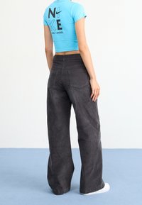 Personne portant un t-shirt Nike turquoise court et un pantalon large taille haute gris foncé, se tenant sur un sol bleu clair contre un mur blanc.