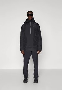 Mann mit schwarzer Kapuzenjacke mit Reißverschluss, schwarzen Hosen, schwarzen Sneakern und großen schwarzen Sonnenbrillen, der vor einem schlichten Hintergrund steht.