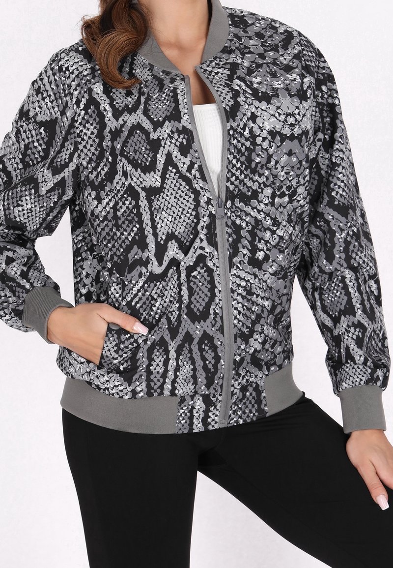 Femme portant une veste bomber zippée noire et grise à imprimé serpent avec poignets et ourlet côtelés, associée à un pantalon noir et un haut blanc.