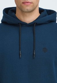 Marineblå hettegenser med snor, med en liten svart logo på brystet. Myk tekstur og standard passform.