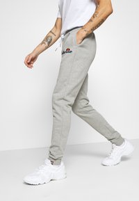 Grå sweatpants lavet af blødt stof med en afslappet pasform, udstyret med sidelommer og et logo på venstre lår. Parret med hvide sneakers.