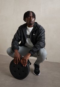 Svart zip-up jacka, vit t-shirt med fet logotyp, grå träningsbyxor, svart viktplatta och svarta sneakers på en strukturerad yta.