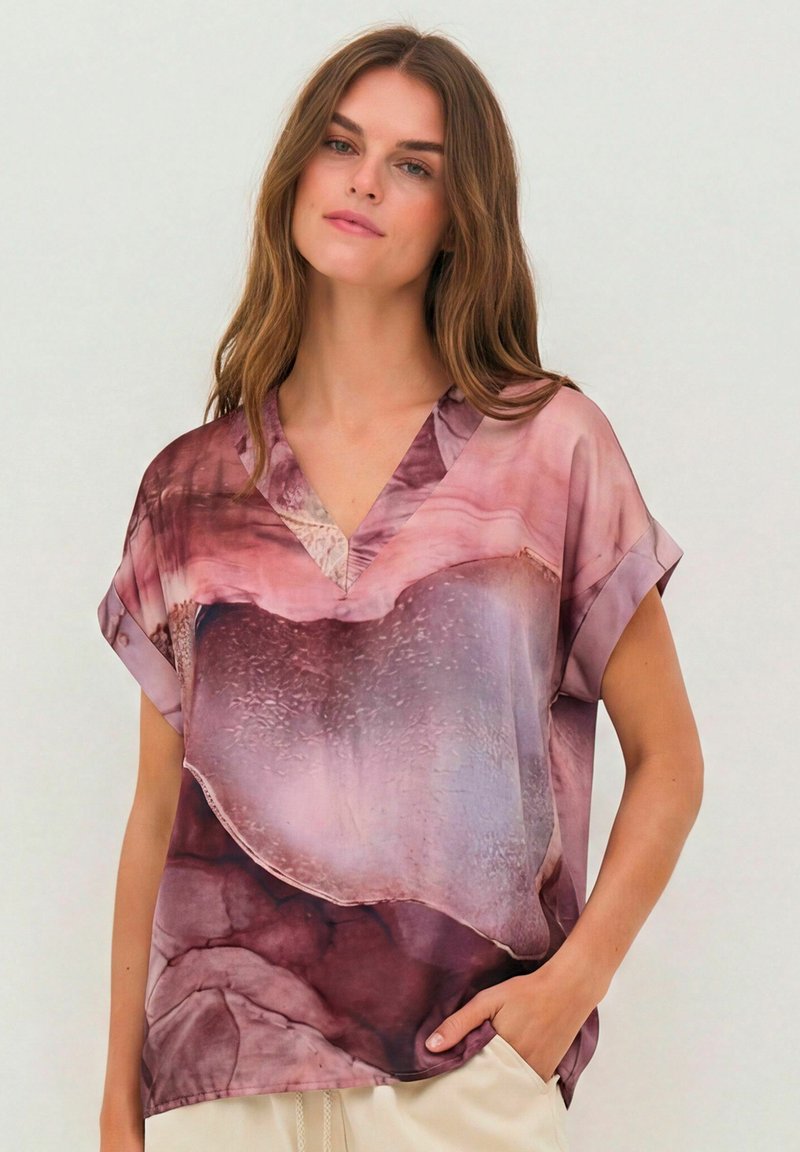 Femme aux longs cheveux bruns portant un chemisier ample à manches courtes en V avec un motif marbré rose et violet, main dans la poche.
