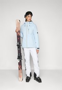 Gaiši zila ūdensizturīga slēpošanas jaka ar rāvējslēdzēju, baltas slēpošanas bikses ar melniem akcentiem, tur rokās slēpes. Valkā melnus brillus un slēpošanas zābakus.