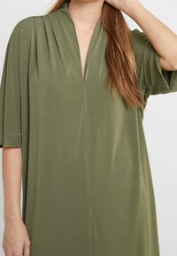 Olivgrön tunikaklänning med djup V-ringning, korta ärmar och mjukt tyg med vertikala veck och minimala sömnadsdetaljer.