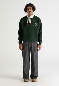 Hombre con cabello rizado y bigote, vistiendo un suéter verde oscuro con logotipo, bufanda de cuello estampada, pantalones anchos de rayas grises, y zapatos color beige, de pie.