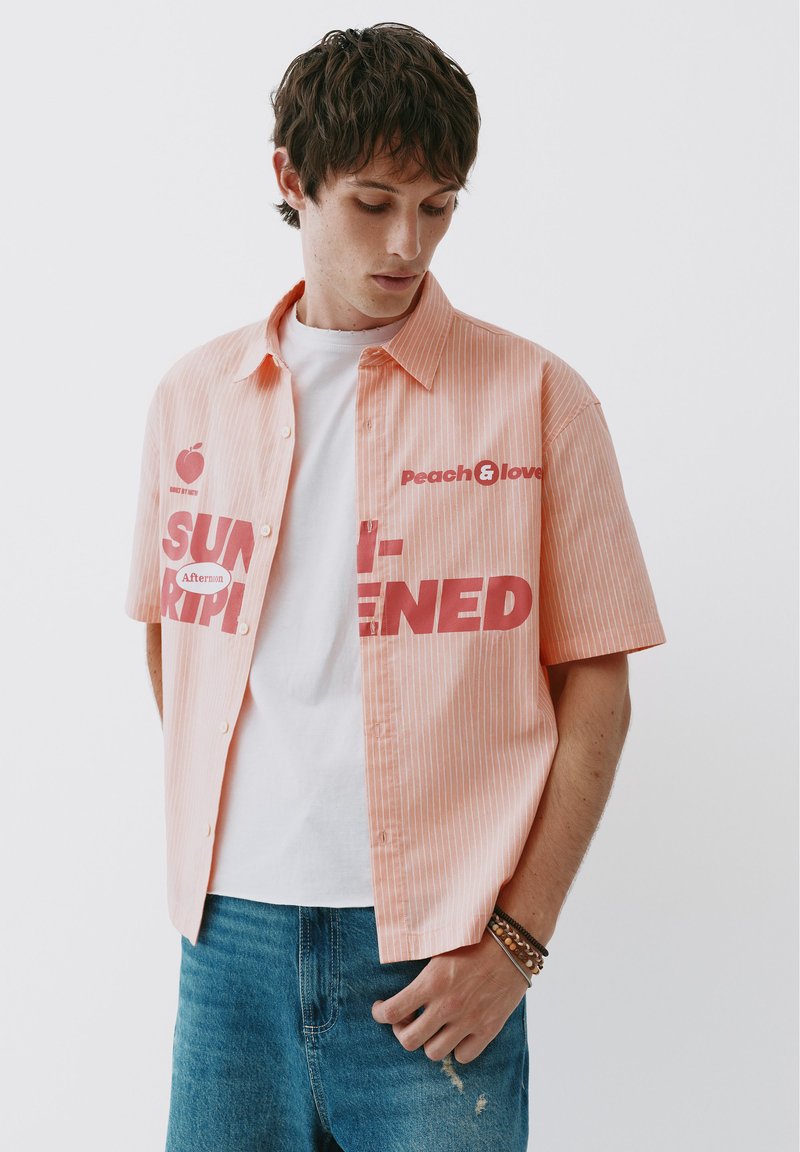 Jeune homme portant une chemise à manches courtes orange clair avec un texte rouge sur un t-shirt blanc et un jean bleu, regardant vers le bas.