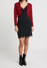 Woman wearing a black lace-trimmed slip dress, red cropped cardigan, and black ankle boots standing against a plain background.