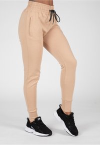 Beige joggerbroek met een trekkoord in de taille, zijzakken en aansluitende boorden, gecombineerd met zwarte sportschoenen met witte zolen.