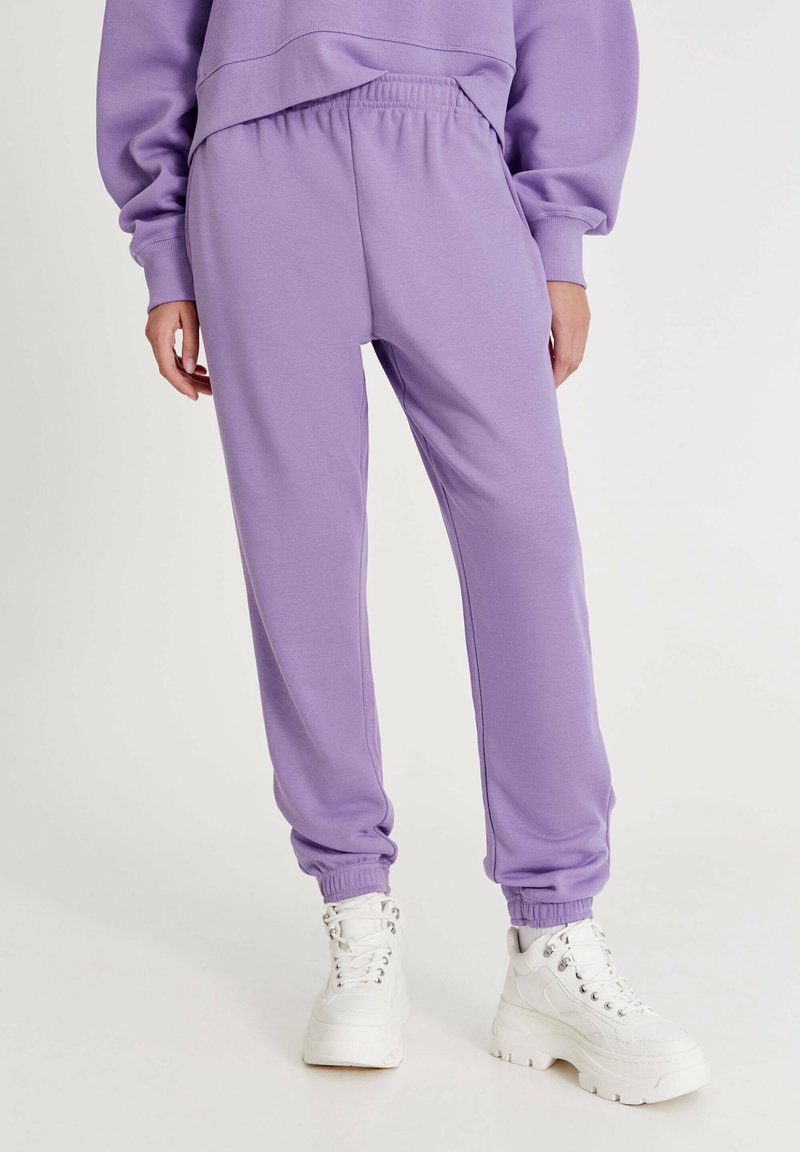 Lilla sweatpants med elastisk talje og manchetter, fremstillet af blødt materiale, parret med hvide chunky sneakers. Glat tekstur, minimal detaljering.
