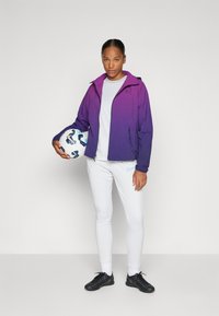 Lilla gradient jakke med hette, grå skjorte, hvite bukser og svarte fotballsko. Holder en mønstret fotball.