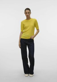 Geel gebreid sweatshirt met korte mouwen, voorzien van een ribbelstructuur en verticale patronen, gecombineerd met donkerblauwe flared jeans en beige schoenen.
