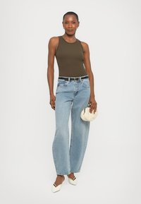Donna che indossa un top senza maniche verde oliva, jeans blu chiaro, scarpe bianche a punta, con una piccola borsa bianca, in piedi su uno sfondo bianco.