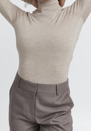 Personne portant un pull à col roulé beige et un pantalon gris taille haute, les mains levées derrière la tête.