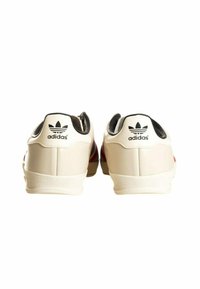 Fehér bőrsneakerek fekete részletekkel, piros és kék csíkos díszítéssel, gumitalppal és jellegzetes Adidas logóval a saroknál.