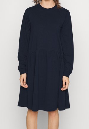 Vrouw draagt een lange mouw, knielange zwarte jurk met een geplooide taille, staand tegen een eenvoudige lichte achtergrond.