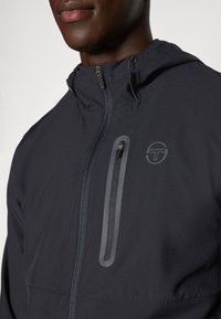 Tumedat meresinise kapuutsiga jope, millel on tõmblukuga rinna-tasku, sujuva tekstuuri ja diskreetse logoga vasakul üleval.