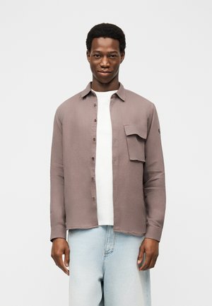 Mees, kes kannab heledat lilla nööbitavat särki valge T-särgi ja helesiniste teksade peal, seisab tavalise heleda tausta ees.