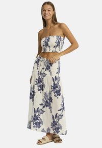 Una giovane donna sorride indossando un crop top floreale senza maniche e una lunga gonna abbinata, con sandali leggeri, su uno sfondo semplice.