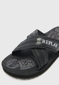 Black flip-flop with fabric straps, featuring a textured footbed and embossed branding. Notable stitching detail on the straps.
