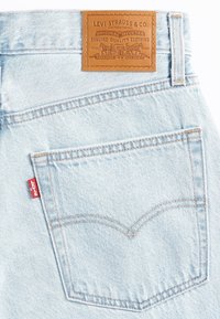 Ljusblå denimjeans har en brun läderlapp med prägling av logotyp, röd märkesskylt på fickan och en klassisk sömnadsdesign för bakfickorna.