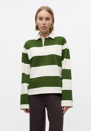 Giovane che indossa una maglia da rugby a maniche lunghe a righe larghe verdi e bianche con pantaloni scuri, in piedi davanti a uno sfondo bianco uniforme.