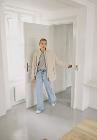 Naine, kes kannab beežist tikitud jopet ja helesiniseid teksaseid, avab valget sisust uksi heledas, minimalistlikus ruumis.