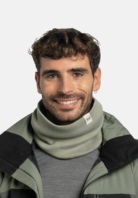 Gaiter de cuello verde claro hecho de tela de punto, con una etiqueta de marca y un diseño ajustado y doblado, usado por un hombre sonriente con ropa de exterior.
