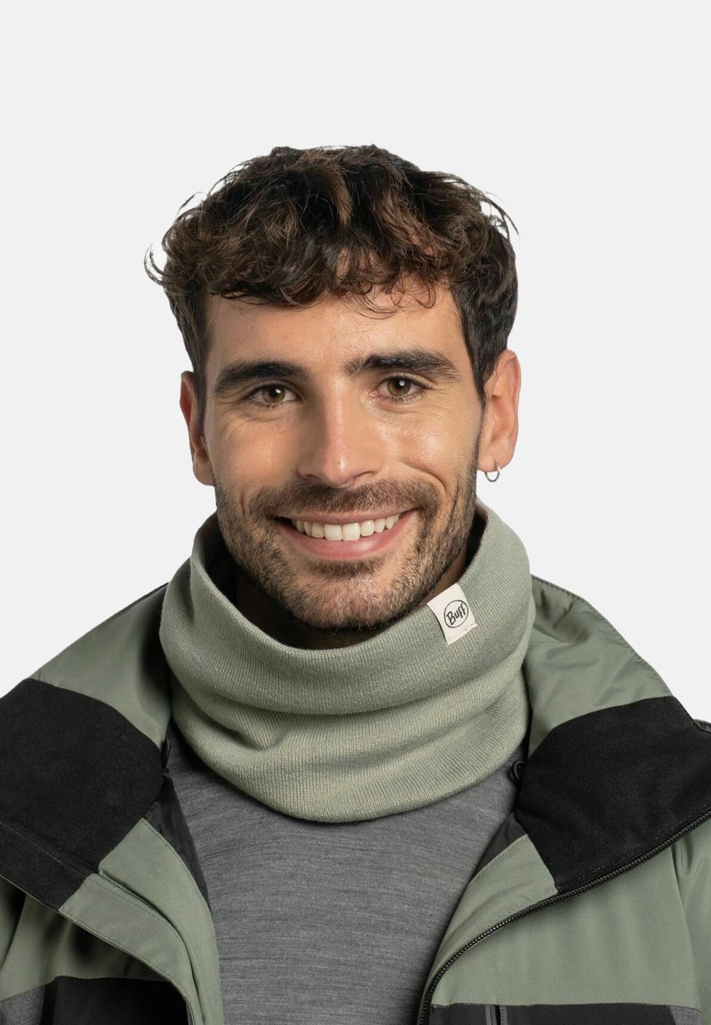 Gaiter de cuello verde claro hecho de tela de punto, con una etiqueta de marca y un diseño ajustado y doblado, usado por un hombre sonriente con ropa de exterior.