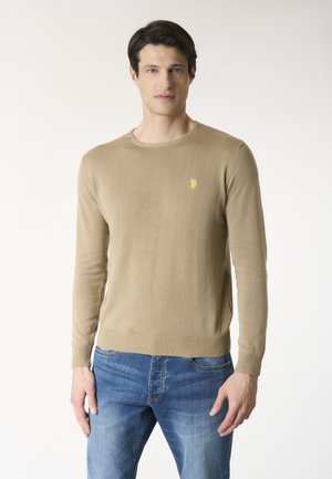 Uomo che indossa un maglione beige girocollo con un piccolo logo giallo sul petto e jeans blu, in piedi davanti a uno sfondo chiaro e uniforme.