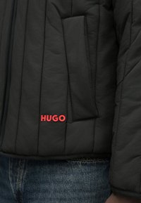 Black quilted jacket with a zip, featuring a left side pocket and red "HUGO" branding on the front. Textured fabric with visible stitching.