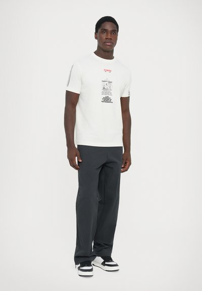 Camiseta blanca de manga corta con texto gráfico en negro y rojo. Combinada con pantalones negros y zapatillas en dos tonos. Diseño sencillo.