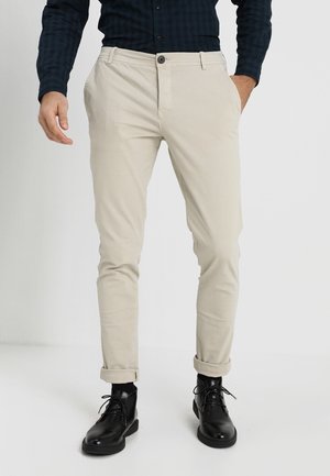 Mand iført beige slim-fit bukser, opsmøgede ved anklerne, sorte læderstøvler med snørebånd og en mørkeblå langærmet skjorte med knapper ved ærmerne.