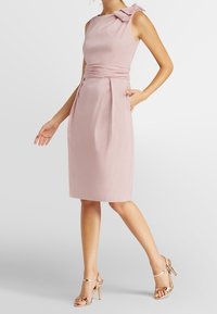 Robe sans manches rose avec un accent en nœud sur l'épaule, corsage ajusté et taille plissée, associée à des sandales à talons dorés avec des lanières.