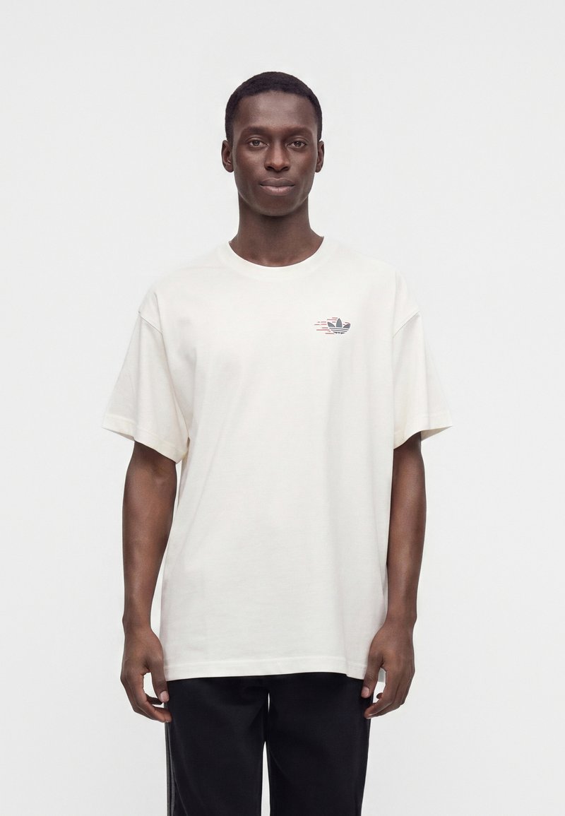 Uomo che indossa una t-shirt bianca semplice con un piccolo logo Adidas sul petto sinistro, in piedi davanti a uno sfondo bianco.
