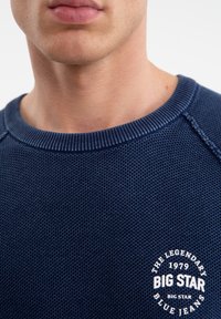 Námorně modrý pletený svetr s texturovaným povrchem. Obsahuje žebrovaný kulatý výstřih a kulatý bílý logo na dolním levém okraji.