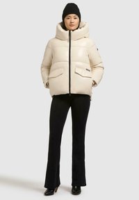 Giacca beige imbottita con colletto alto, zip frontale e due tasche frontali; indossata sopra pantaloni neri aderenti e stivaletti.