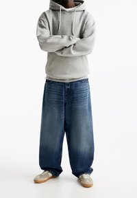 Sweat à capuche gris, jean bleu foncé ample, et baskets grises avec des accents clairs. Coupe large et style décontracté.