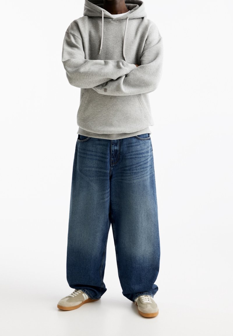 Sweat à capuche gris, jean bleu foncé ample, et baskets grises avec des accents clairs. Coupe large et style décontracté.