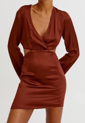 Femme portant une mini-robe en satin marron, cintrée, à manches longues, col en V profond avec des découpes sur les côtés, debout contre un fond uni.
