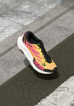 Sapatilha de corrida em amarelo brilhante e rosa, com atacadores pretos, sola branca e a marca "HOKA", sobre pavimento urbano texturizado perto do meio-fio.