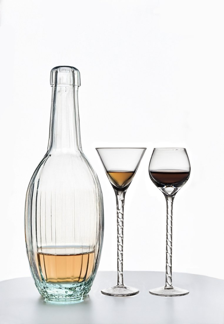 Durchsichtige Glasflasche mit ribbeligem Design, gefüllt mit hellem Bernsteinflüssigkeit. Zwei klare Stielgläser: eines in Martinitasse-Form, das andere kurz und rund, jedes mit brauner Flüssigkeit.