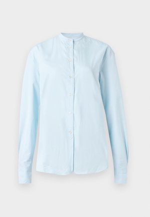 Γυναίκα στέκεται με τα χέρια στις τσέπες φορώντας ένα ανοιχτομπλε πουκάμισο με κουμπιά, φαρδιά μαύρη παντελόνα και μαύρες σαγιονάρες σε λευκό φόντο.